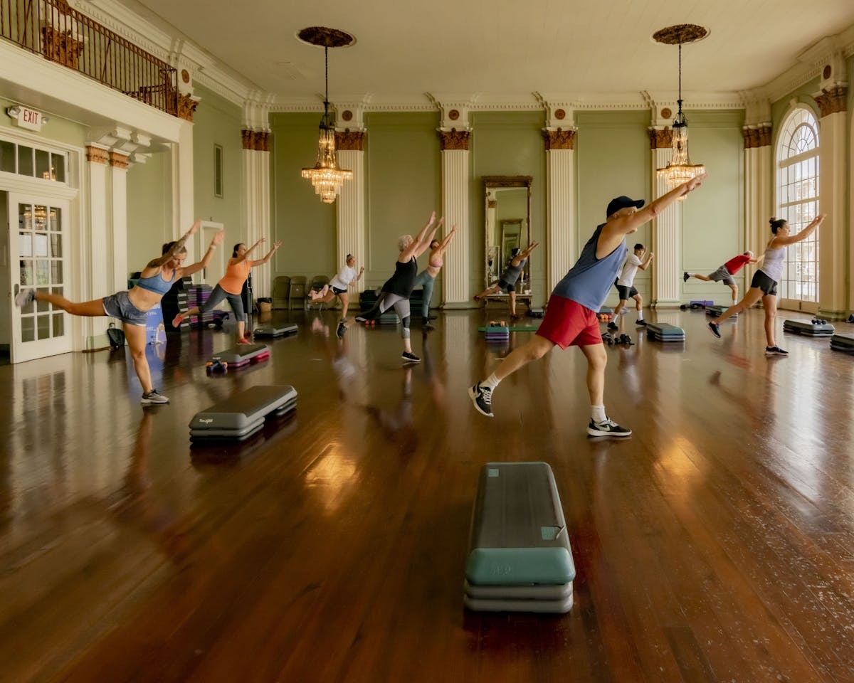 Group of people participating in a fitness class in a large room. The room has a wooden floor, large side windows, and chandeliers.
