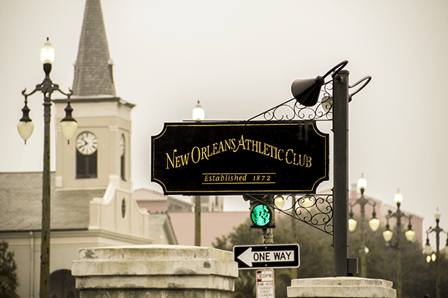 A black sign hanging from a black wrought iron post that reads “New Orleans Athletic Club” in gold lettering, with “Established 1872” below it.