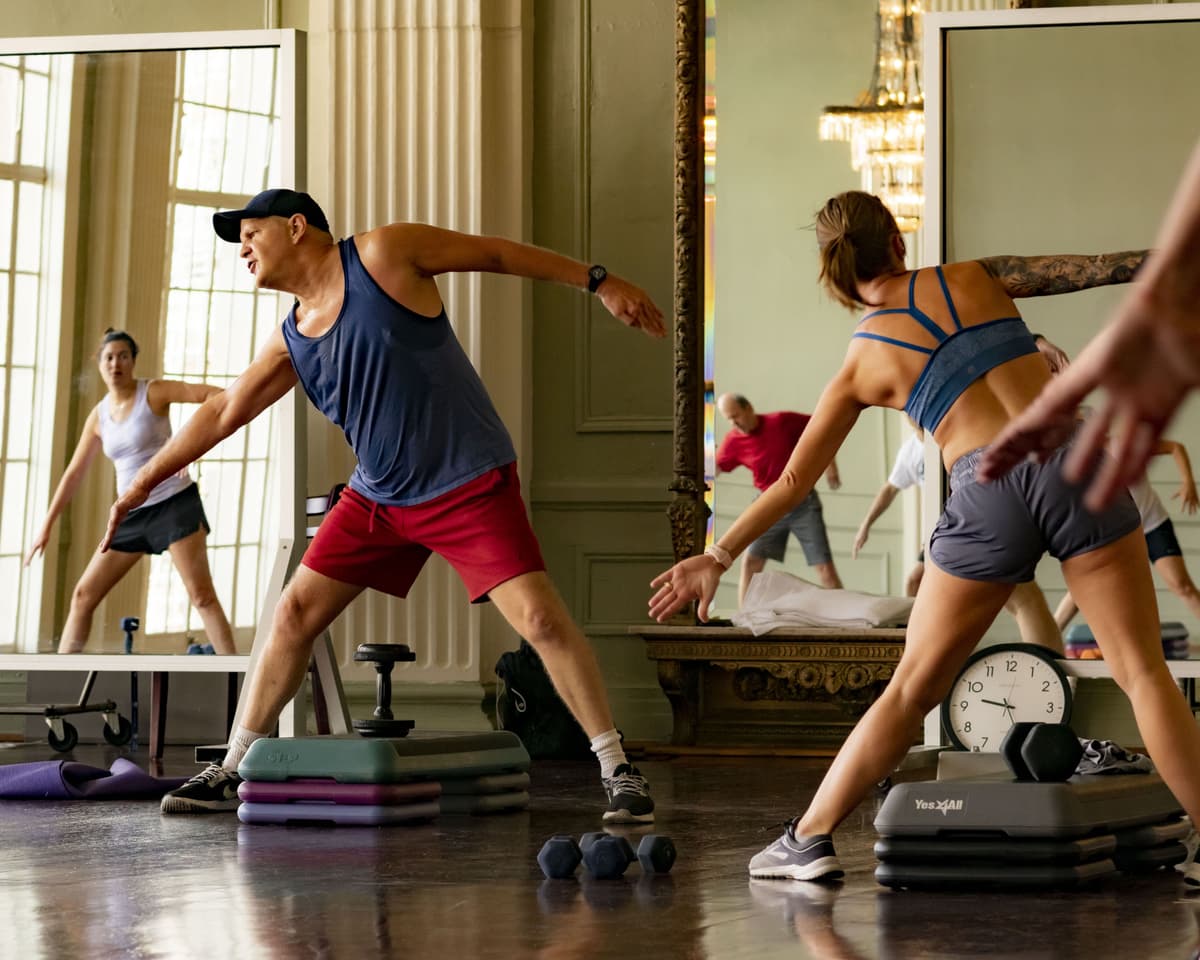 A gym instructor led a group of people in their fitness session. Various equipment on the floor includes step platforms between their legs, mats, and weights.