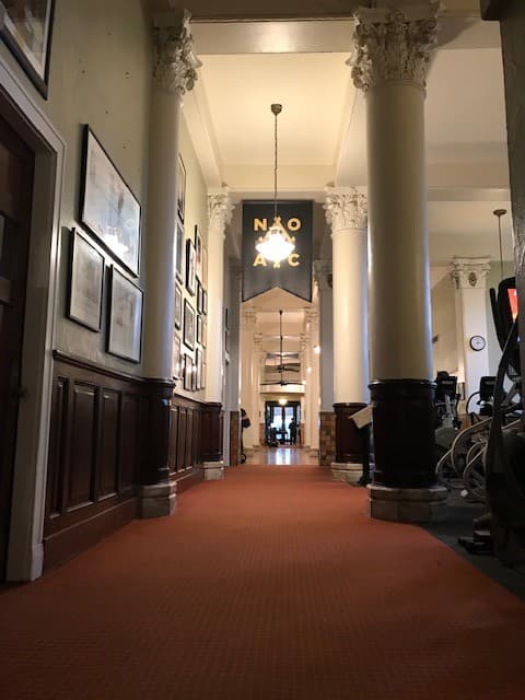 The gym hallway is painted white and has frames on the wall. In the center hangs a black banner with the NOAC logo.