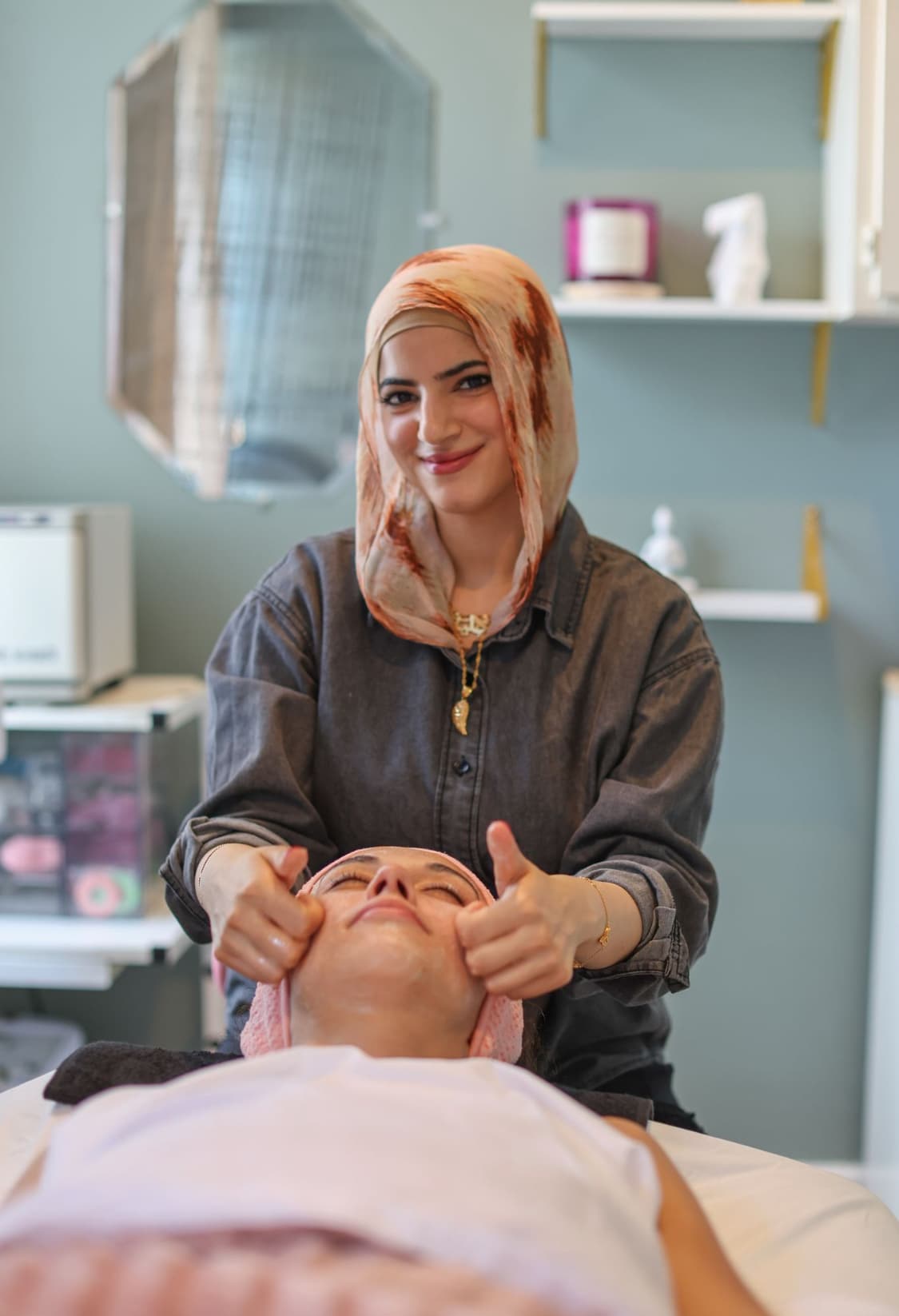 A nail salon has two white pedicure chairs, beige towels, and a black rolling chair in front of the chairs. It has a white shelf with nail polish bottles between the two chairs.
