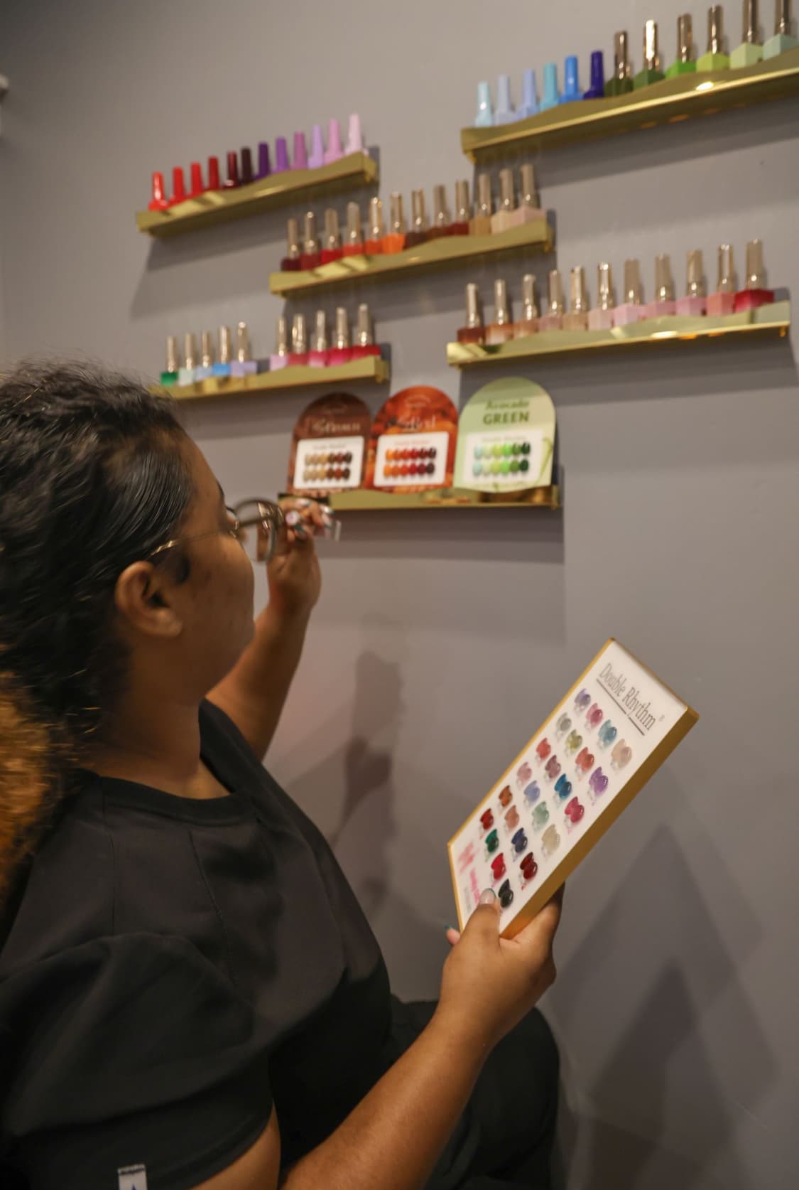 A nail salon has two white pedicure chairs, beige towels, and a black rolling chair in front of the chairs. It has a white shelf with nail polish bottles between the two chairs.