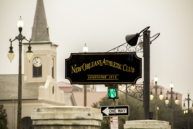 A black sign hanging from a black wrought iron post that reads “New Orleans Athletic Club” in gold lettering, with “Established 1872” below it.