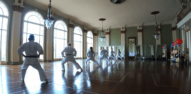 A group of people is practicing karate stances in a gym room. The room has a wooden floor, big arch windows, and chandeliers.
