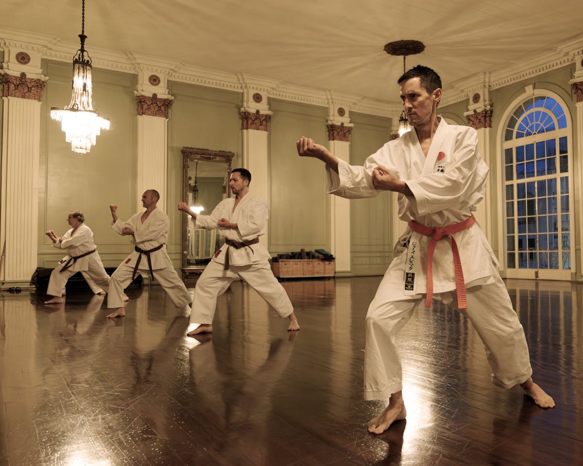 A group of people is practicing karate stances in a gym room. The room has a wooden floor, big arch windows, and chandeliers.