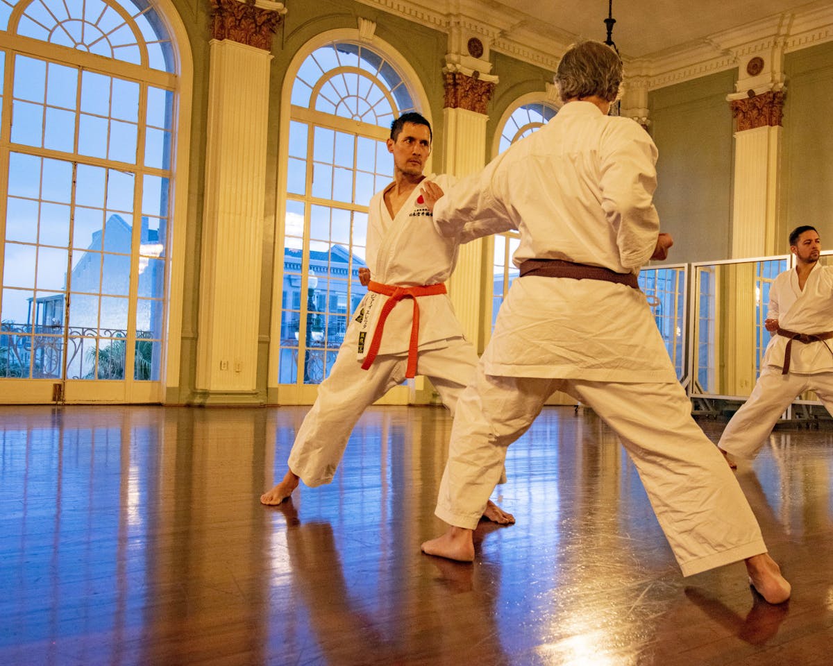 The instructor and trainee have one-on-one karate practice in the gym. They are inside a large room with a wooden floor and high arch windows.
