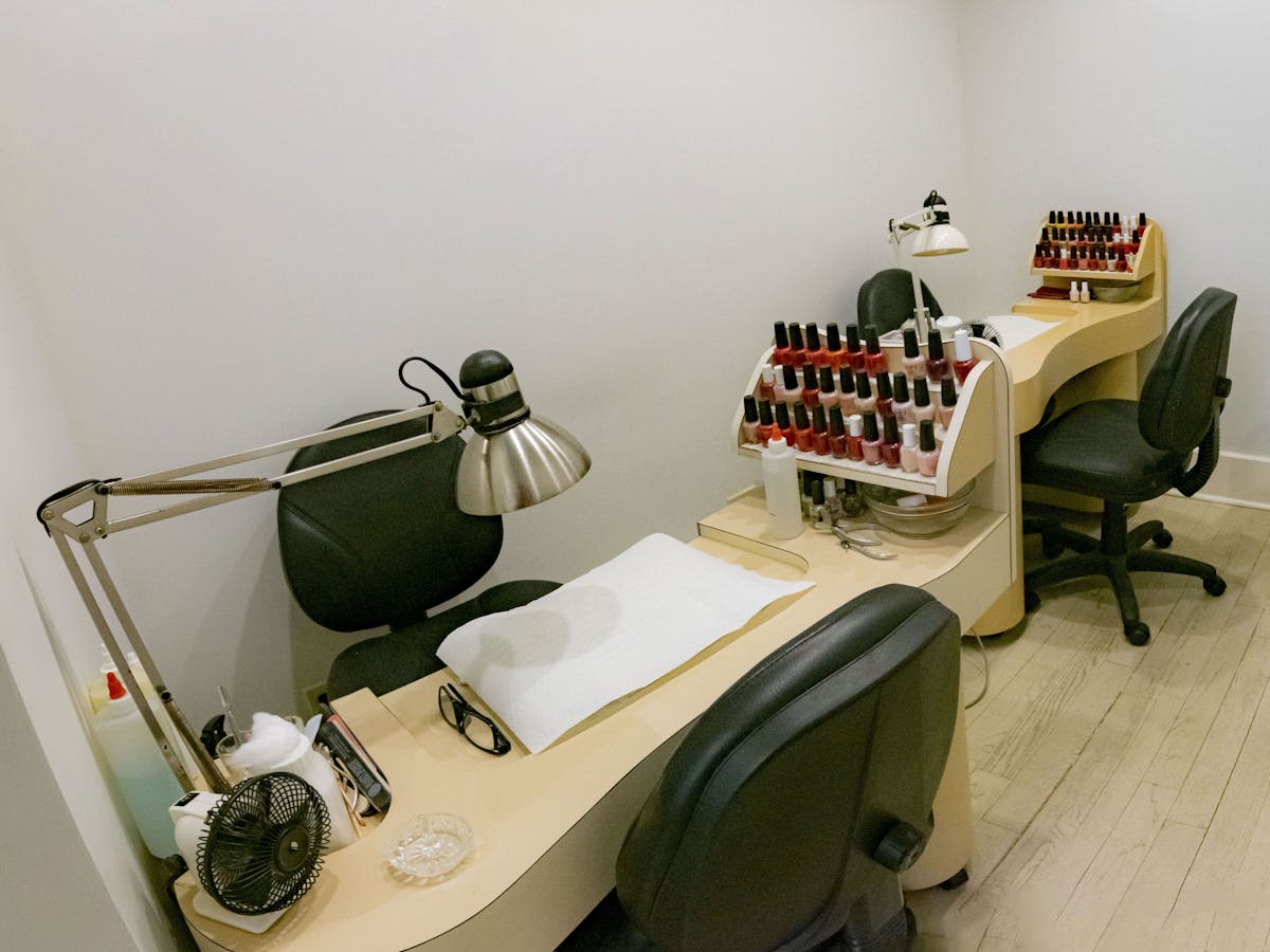 A nail salon station with two chairs and a desk. The desk has a lamp, a fan, and a rack of nail polish bottles.