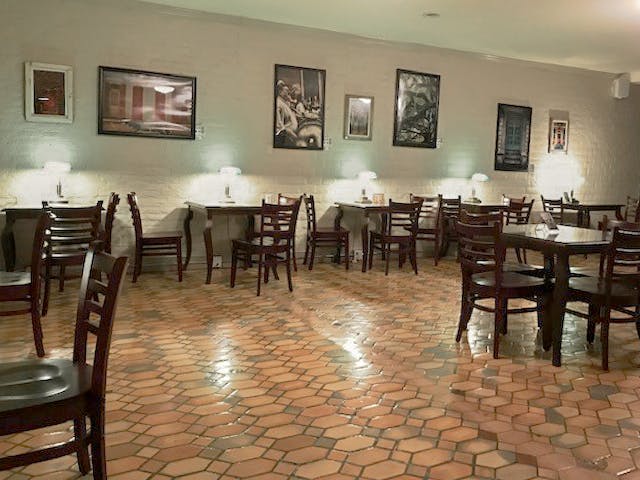 A bar interior has hexagonal tiles and white walls with several frames. There are several wooden tables and chairs with green lamps on the tables.