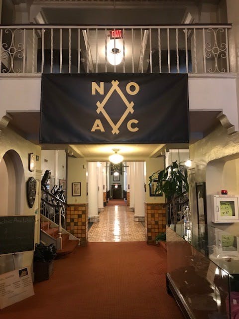A hallway with a large black banner hanging from the ceiling. The banner has gold lettering and symbols that read “NOAC.” 
