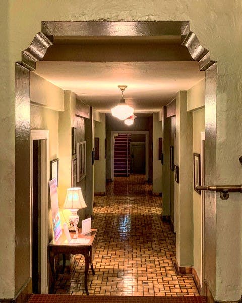 A hallway with a  brick floor and white colored walls with frames attached to it. It has a table with a lamp on the left side.