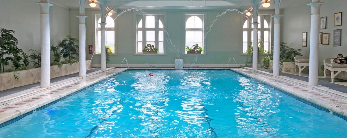 Indoor rectangular-shaped swimming pool with a person swimming in it. White columns and arches surround it, and two fountains are on either side of the pool.