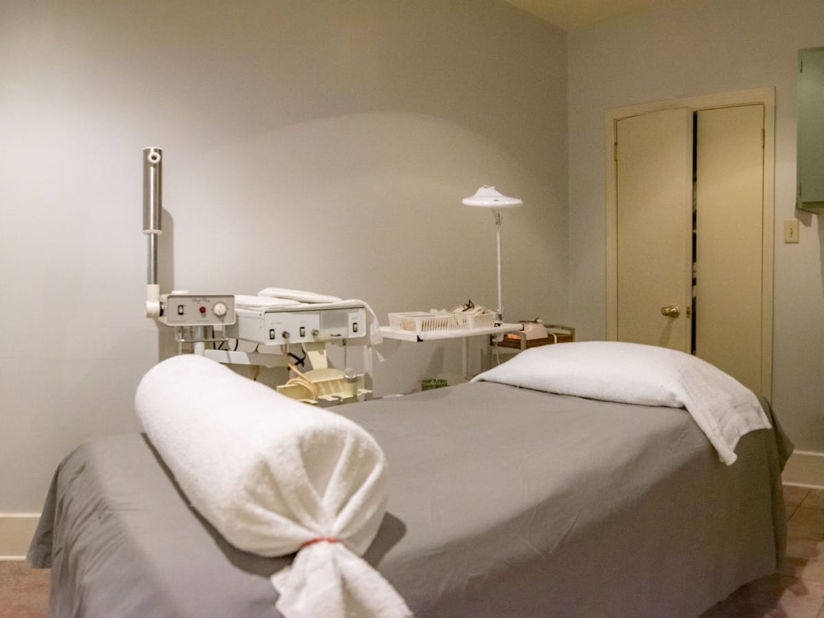 A massage room with a massage table in the center. It is covered in a gray sheet with white towels on top. On the left side are various equipment and materials.