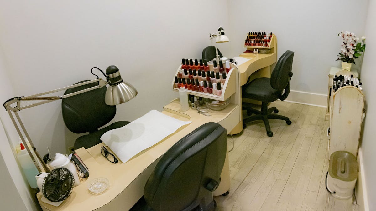 A nail salon station with two chairs and a desk for manicures. The desk has a lamp, a fan, and a rack of nail polish bottles.