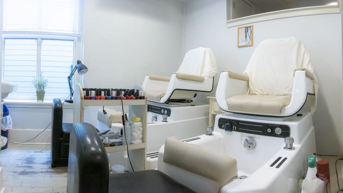 A nail salon has two white pedicure chairs, beige towels, and a black rolling chair in front of the chairs. It has a white shelf with nail polish bottles between the two chairs.